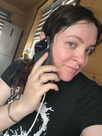 A medium-shot, angled, indoor portrait captures a woman holding a black smartphone to her left ear. She appears to be on a call, and a white charging cable is connected to the phone, visible near her wrist. She is wearing a black t-shirt with a white graphic print. A gold-colored bracelet adorns her left wrist, and a multi-stone ring is on her ring finger. The woman has light skin, dark hair, and blue-grey eyes. The background includes what appears to be a wooden wall, a window, and part of a refrigerator. Specific location details regarding Protasy, Russia are not discernible from the image.
