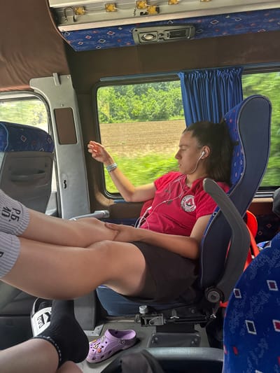 A young woman with dark hair tied in a ponytail, wearing a red t-shirt and black shorts, sits in a bus seat, looking out the window. She has white earbuds in her ears and a bracelet on her wrist. Her legs are propped up, with one foot wearing a grey and white striped sock and the other foot out of frame. In the foreground, another person's feet are visible, one wearing a black sock and a white slide, and next to it, a purple Croc shoe with charms. The bus interior features blue seats with a diamond pattern, brown upholstery, and blue curtains. Outside the window, a field and trees are visible, indicating a rural or semi-rural setting, potentially near Izvorovo, Bulgaria, based on context. The lighting suggests it is daytime.