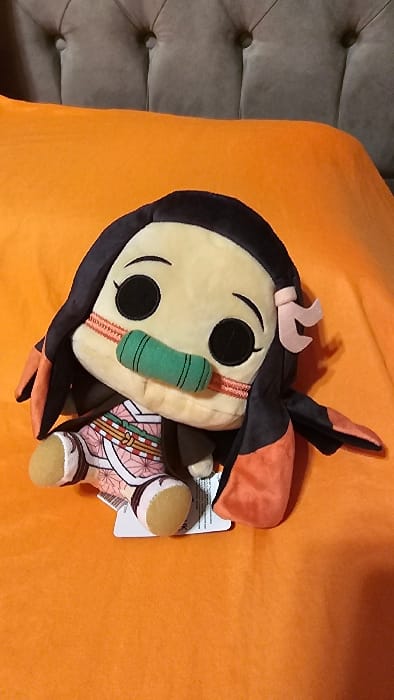 A Nezuko Kamado plush doll lies on an orange surface with a gray tufted pillow in the corner. The doll features a tan face with large black eyes and a green bamboo muzzle. Pink accessories adorn the black hair. The doll wears a pink kimono with a white and brown geometric pattern. The doll is on its side with a white tag still attached to one of its legs.