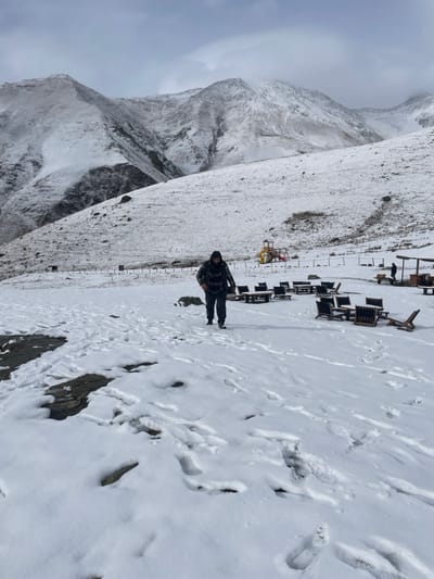 The image depicts a snow-covered mountainous landscape in the region near Juta, Georgia, under an overcast sky. In the foreground, snow covers the uneven ground, revealing numerous footprints and sporadic patches of darker rock. An adult male, dressed in a dark hooded jacket and dark trousers, walks from the left towards the right across the mid-ground, leaving discernible footprints in the snow.

Behind the walking individual, a fenced area contains multiple sets of dark-colored outdoor tables and chairs, arranged on the snow. To the right of these furnishings, a brightly colored children's playground structure, primarily yellow, red, and blue, is visible. Further to the right, near a small wooden shelter, two indistinct human figures are present. The background features steep, snow-covered mountains with rocky outcrops, partially obscured by low clouds or mist. The overall environment indicates cold, snowy conditions.