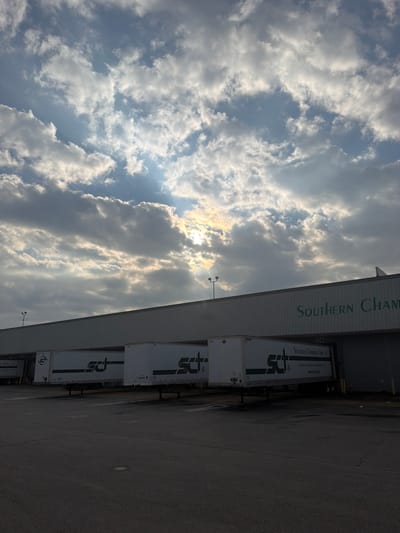 The image depicts the exterior of a commercial building, likely a warehouse or distribution center, in Chattanooga, Tennessee, United States. The building has a long, grey, corrugated metal facade. The name "SOUTHERN CHAMPION" is visible in large green lettering across the top of the building. Parked adjacent to the building are several white semi-trailer trucks, some bearing the logo "SCI" in green. The trailers are positioned at loading docks, indicated by the raised platforms and doors. The ground in front of the building is paved asphalt. Above the building, the sky is filled with dramatic clouds, with sunlight breaking through in several areas, creating a partly cloudy and somewhat dramatic sky. The overall scene suggests a working industrial or commercial environment under a dynamic sky.
