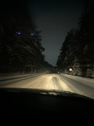 A nighttime shot from inside a vehicle shows a snow-covered road stretching forward between snow-laden trees. The road is illuminated by headlights, creating a bright path through the darkness. A road sign is visible further down the road. The sky is dark, and the edges of the frame are obscured in shadows, focusing the viewer's attention on the illuminated road ahead. Snow is heavily piled on the sides of the road and on the trees creating a tunnel effect.