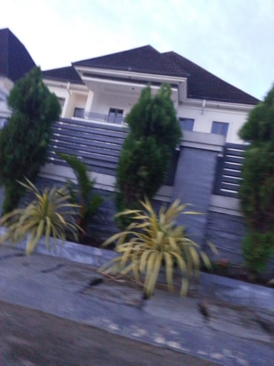 The image shows a contemporary two-story house with a dark, sloped roof, white exterior walls, and dark window frames. The house features balconies with metal railings and a modern, horizontal fence with dark and white elements. In front of the house, there are several large, green, conical trees and a bed of low-lying, yellow-green plants. The foreground is dominated by a dark, textured surface, possibly a road or pathway, with some visible horizontal lines. The sky is overcast and pale. The overall scene suggests a residential property in a suburban or developed area.