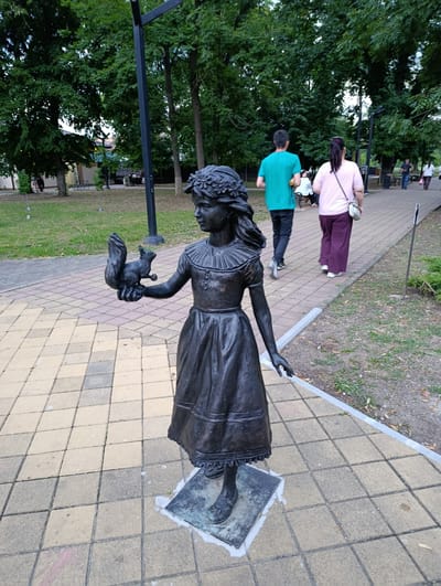 In Kelermesskaya, Russia, a bronze statue of a young girl wearing a floral wreath and a dark dress stands on a paved walkway. She is holding a squirrel in her outstretched left hand. In the background, several people are walking on the same path. One person in a teal shirt and dark pants walks away from the camera, while a woman in a pink shirt and purple pants walks ahead. Trees line the walkway, and a park with grass and benches is visible to the left. The lighting suggests it is daytime.