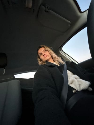A person with long blond hair sits in a car, looking up toward the camera. They wear a black coat over a white shirt or sweater. A seatbelt crosses their chest. The interior of the car is dark, with the sunroof visible in the top left corner. The car's sun visor is also visible. The camera angle is from below, looking up toward the subject.
