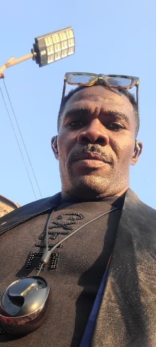 The image displays an adult male from a low-angle perspective, located in the City of Alayi, Nigeria. The subject has dark skin, short dark hair, a mustache, and a short beard. A pair of light-framed eyeglasses rests on top of his head, and an in-ear device is visible in his right ear. He wears a dark t-shirt with a dark, textured pattern on the chest, overlaid by a dark, reflective jacket. A polished, metallic, oval-shaped pendant is suspended around his neck by a dark cord. The background consists of a clear blue sky. In the upper left, a multi-panel LED street light is mounted on a pole, with a thin dark wire extending from the lower left towards it. A faint, light-colored structure is partially visible in the far upper left corner of the frame. The scene is illuminated by direct sunlight.