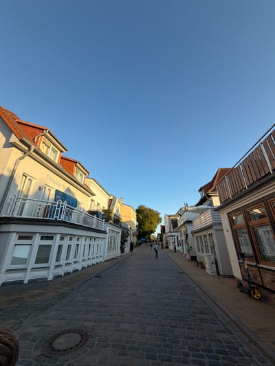 The image depicts a cobblestone street lined with buildings on both sides, under a clear blue sky. The street appears to be in a town or city, with a few people walking along it. On the left side, a building with a sign reading "Pension StromInn" is visible. The architecture suggests a European style, possibly coastal given the presence of beach chairs on a balcony. On the right side, there are more buildings with wooden railings and windows. A scooter is parked near one of the buildings on the right. The overall scene is one of a pleasant, sunny day in a well-maintained urban or town environment. The location has been identified as the Mönchstraße in Rostock, Germany.