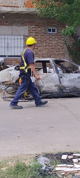 This daytime image, taken in San Fernando, Argentina, shows a man in work attire standing next to a severely damaged, burned-out vehicle. The man is wearing a yellow hard hat, a blue short-sleeved shirt with yellow reflective stripes, blue trousers, and a safety harness. He is holding a clear plastic bag or container in his left hand, which appears to contain wires or other small items, suggesting an activity related to inspection, collection, or salvage.

The vehicle, likely a car or truck, is extensively charred and stripped, with only its metal frame and some distorted body panels remaining. It is covered in soot and appears to have been subjected to intense heat.

In the background to the left, there is a brick building with a small, white-framed window. Green tree foliage hangs over the top part of the building. The ground is a paved surface, possibly concrete or asphalt, leading towards a grassy verge on the right side of the frame. This verge is strewn with debris, including broken tiles and a white plastic bag. The overall scene suggests the aftermath of a vehicle fire, with a worker or investigator on site. There is no visible text in the image.