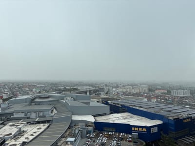 This is an aerial, high-angle, medium shot of a hazy, overcast urban landscape in Sao Thong Hin Town Municipality, Thailand. The foreground is dominated by the rooftops of a large complex of modern buildings. Prominently featured is a bright blue IKEA store with its distinctive yellow logo, its roof covered in a grid of solar panels. Adjacent to the IKEA store is another large, grey, multi-level building with a curved facade. The foreground also includes a parking lot filled with many cars, predominantly white and black, and a complex system of rooftop ventilation and mechanical equipment. In the midground and background, the city unfolds with a dense collection of smaller buildings, residential homes with red-tiled roofs, and a distant cityscape partially obscured by the pervasive haze. The sky is a uniform, light grey, suggesting heavy cloud cover or smog.