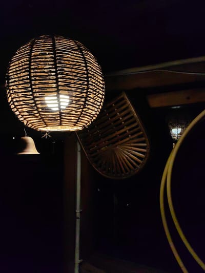 The image captures an outdoor scene at night, likely in Aleo, India, illuminated by two woven pendant lights. The primary light source is a large, spherical woven lamp made of natural fibers, emitting a warm glow from within. Hanging to the left of this lamp is a small, bell-shaped object. In the background, a second, smaller woven light fixture is visible, also lit from within. To the right, a yellow, curved pipe or hose is prominent in the foreground. A decorative woven fan-like object is positioned behind the main lamp. The overall atmosphere is dark and ambient, suggesting an evening setting.