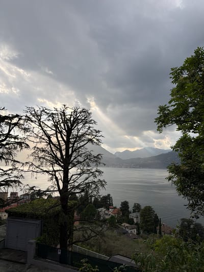 The image captures a view from Gittana, Italy, overlooking Lake Como. The sky is overcast with grey, heavy clouds and beams of sunlight piercing through. Silhouette of a large, leafless tree dominates the foreground, partially obscuring the lake and the distant mountains on the opposite shore. Residential buildings are visible along the shoreline. Green foliage appears to be framing the view from the top of the image. A small grey shed and a fence are visible at the bottom right. The landscape is viewed from an elevated position.