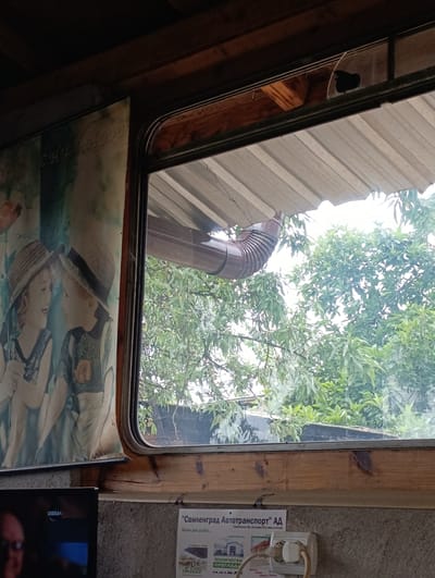 The image shows an interior view looking out a window. On the left, a poster depicting two children in straw hats is partially visible. The poster has the word "CHILDHOOD" written across the top in a stylized font. The window frames an outdoor scene with lush green foliage and a brown downspout visible against a light sky. Below the window, a television screen with the brand name "DIEMA" is visible on the left. On the right, a white electrical outlet with a plug is mounted on a textured wall, and a flyer is attached to the wall below it. The flyer contains text in Cyrillic script, including the phrase "Свиленград Автотранспорт" (Svilengrad Autotransport). The overall impression is a rustic or older indoor setting with a view of nature.