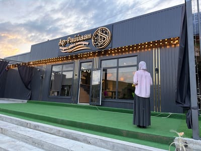 This is a medium-shot, exterior, eye-level photograph taken at dusk on a cloudy day, showing the front of a modern establishment identified by Arabic and Cyrillic signage as "Ар-Райяан" (Ar-Rayyan), with additional text "кофе & еда & эмоции" (coffee & food & emotions). The building features a dark, ribbed metal facade with a decorative band of warm-toned string lights running along its upper edge. A large, circular, gold-colored emblem is mounted above the entrance.

The entrance itself is partially open, revealing a glimpse of the interior. Multiple large windows offer views into the dining area, where tables, chairs, and lighting fixtures are visible. A ceiling fan is noticeable through one of the windows.

In front of the building, a tiered area is covered with artificial green turf. A person, viewed from behind, is standing on the turf wearing a light pink, striped long-sleeved shirt and a long, dark gray pleated skirt. They are facing away from the camera, towards the building.

To the left of the building, a black fabric canopy or curtain is partially deployed, and to the right, a black flag or curtain is hanging. Concrete steps lead up to the turf area from the foreground. The overall environment suggests an outdoor seating or waiting area for a cafe or restaurant. The city of Tereze, Russia is not visually discernible from the image.