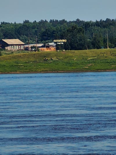 The image shows a wide, calm body of water in the foreground. In the midground, a grassy embankment slopes upwards towards a settlement. On the embankment, there is a cluster of buildings, including a traditional wooden house with a light-colored roof and several windows. To the right of the house, there are some smaller structures and what appear to be stacks of lumber or wood. Above the settlement, a sign with Cyrillic lettering that reads "ПОТАПОВО 1670" is visible, indicating the location is Potapovo, Russia, and that the settlement dates back to 1670. Behind the settlement, a dense line of green trees forms the background under a clear, pale blue sky. The overall scene depicts a rural settlement situated along a river.