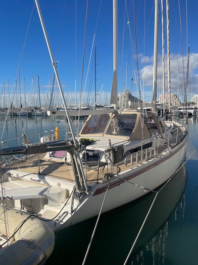 The image depicts a marina in La Grande-Motte, France, under a clear blue sky with direct sunlight. In the foreground, a white sailboat with a dark red hull stripe is moored in calm, dark blue water, reflecting its hull. The boat features a light wooden deck, a tan canvas dodger with clear plastic windows covering the cockpit area, and various ropes and lines coiled on the deck. A two-masted rigging system is partially visible, with white masts extending vertically. An inflatable dinghy, light grey in color, is tethered to the stern of the foreground vessel. In the midground and background, numerous other sailboats are docked, their white masts and rigging forming a dense vertical pattern against the sky. On the distant horizon, several distinctive white, modern, pyramid-shaped buildings, characteristic of La Grande-Motte's architecture, are visible above the other boats. No individuals are present in the frame.