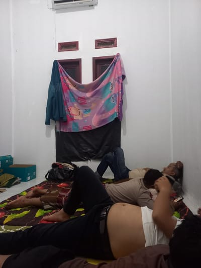 The image shows a dimly lit room with white walls, possibly a bedroom or living space in a residence in Puloblang, Indonesia. Four people are lying down on what appears to be a bed or mattress covered with a patterned sheet. One person in the foreground is wearing a white t-shirt and black pants, with their abdomen visible. Another person is lying beside them, wearing a beige t-shirt and jeans. A third person is partially visible, wearing a brown shirt, and a fourth person is seen from the back, with dark hair. A colorful blanket or sheet with cartoon characters and the text "AIR" and "YOUR" is draped over the people. A dark tarp is spread out behind them. To the left of the frame, there are two small rectangular windows with dark frames and what looks like an air conditioning unit mounted on the wall. On the floor, there are two blue cardboard boxes and some bags. The overall atmosphere seems relaxed, with the individuals resting. There are no clear indicators of the time of day or weather, but the indoor lighting suggests it might be evening or night.