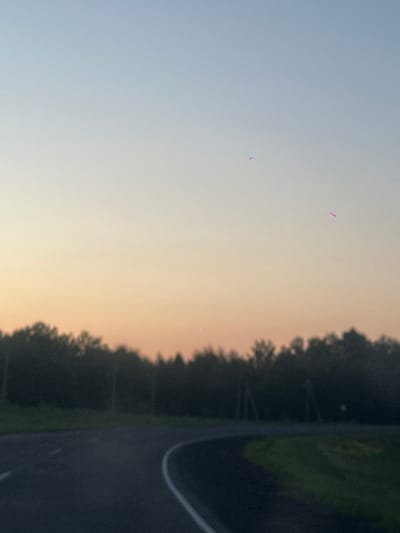 The image depicts a sunset over a rural landscape, likely near Krapivinskiy, Russia. The sky transitions from a light blue at the top to hues of orange and yellow near the horizon. A dark silhouette of a dense forest forms the backdrop against the colorful sky. In the foreground, a paved road curves through the scene, with white lane markings visible. The overall impression is one of tranquility and the end of a day. There are no people or animals visible in the image.