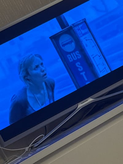 The image shows a television screen displaying a scene from a movie or show. A woman with blonde hair is looking upwards with a surprised or concerned expression. To her right, there is a blue bus stop sign with white lettering. The text on the sign reads "BUS STOP" and "BUS 119". The background is predominantly blue, suggesting an indoor or nighttime setting. The bottom of the image shows the stand of the television, with the "SAMSUNG" logo visible. There are also some white cables connected to the television. The scene appears to be set in a city, as indicated by the bus stop sign. However, there is no information in the image that can definitively identify the specific location as Sovkhoza imeni Lenina, Russia.