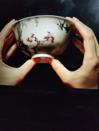 Two hands are holding a white porcelain bowl with floral and bird motifs. The bowl is decorated with pink cherry blossoms and two black birds in flight. The hands are positioned on opposite sides of the bowl, with the fingers curled around its exterior and the thumbs supporting its base. The background is dark, suggesting an indoor setting. The bottom of the image shows text that appears to be a digital interface, with the words "erusia 300 Tahun Ini Laku..." and "Simpan" visible. There is no information available regarding the location in the city of Kualaraja, Indonesia.