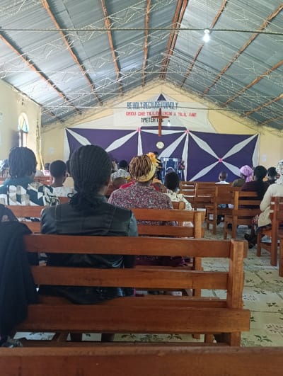 The image captures the interior of a church or meeting hall in Kaplelwo, Kenya, from a rear perspective, looking towards the front. A congregation is seated in wooden pews, mostly viewed from behind, facing the front of the hall.
The main subjects are the attendees, dressed in various casual and semi-formal attire. In the immediate foreground, a person wearing a black leather jacket is seated. Several other individuals are visible further back, including some on a stage or platform at the front.
The setting is a simple structure with exposed corrugated iron roofing and wooden rafters overhead. A single light bulb hangs from the ceiling. The floor is covered with light-colored, patterned tiles. The front of the hall features a decorative display: a large white and purple fabric banner with diamond patterns. Above this, a larger white banner displays text.
The activity taking place appears to be a religious service or community gathering, with people seated attentively, likely listening to a speaker or participating in worship. The general atmosphere seems reverent.
Notable details include the construction style, indicative of a functional and possibly local community building. The time of day appears to be daytime, with some natural light entering, although an artificial light source is also active.
Visible text on the main banner reads: "BE HOLY BECAUSE I AM HOLY" and "1 PETER 1:16". Below this English text, there is also text in another language, likely a local Kenyan language, which says: "DEGU CHE TILILE ANUN A TILIL ANE".
