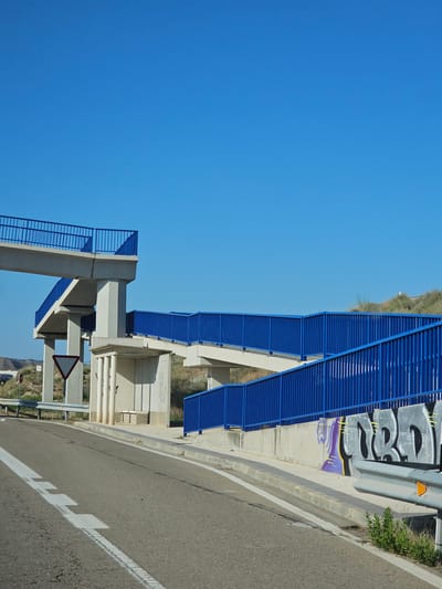 The image captures a pedestrian overpass with bright blue railings, stretching across a road in Zaragoza, Spain. The overpass is supported by sturdy concrete pillars, and a ramp leads up to it. The road surface is asphalt with white lane markings. In the background, a dry, scrubby landscape with some sparse vegetation can be seen under a clear blue sky. There is graffiti on the concrete wall beneath the overpass. No people are visible in the image.