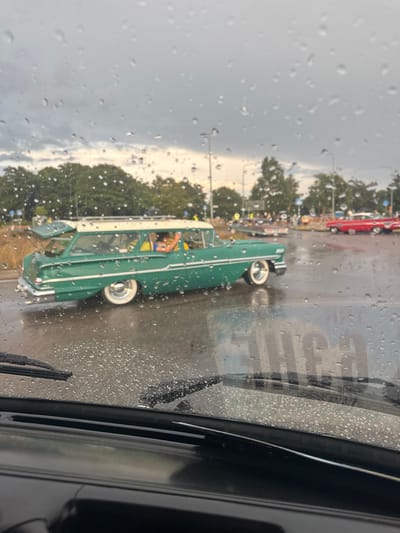 The image shows a rainy day scene, viewed from inside a car, likely in Falkenberg, Sweden. Water droplets are visible on the car's windshield, distorting the view of the outdoor environment. A vintage green station wagon with a white roof is the primary subject in the foreground, driving on a wet road. The car has whitewall tires and chrome accents. In the background, other classic cars, including a red one, are parked or driving, suggesting a car meet or event. Trees and streetlights are present, indicating an urban or suburban setting. The sky is overcast, consistent with rainy weather.