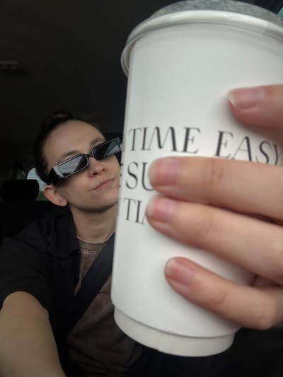 A woman wearing rectangular sunglasses and a seatbelt is holding a white coffee cup that reads "TIME EASY". The photograph appears to be taken from inside a vehicle. The location, Kreshchenye Kazyli, Russia, cannot be confirmed from visual data alone.