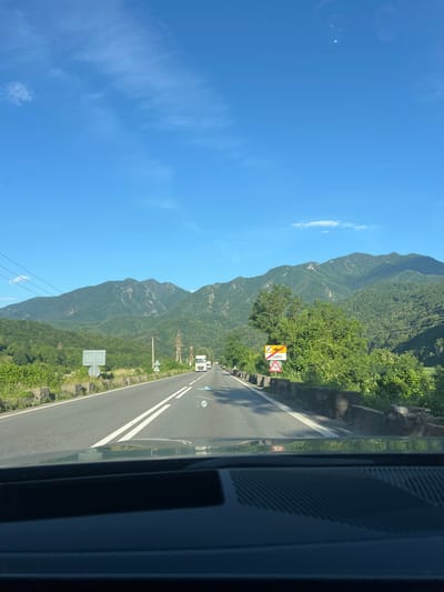 The image depicts a view from inside a vehicle driving on a two-lane road towards mountains under a clear blue sky. The road has white dividing lines and a concrete barrier on the right side. Several trucks are visible on the road ahead. On the right side of the road, there are two traffic signs, one indicating a speed limit of 30 and another with a symbol of a deer, suggesting a potential wildlife crossing area. The text "Fieni" is visible on one of the signs. The background features lush green mountains with some trees and electrical pylons. This scene is consistent with the landscape found in the vicinity of Brezoi, Romania, known for its scenic mountain roads.