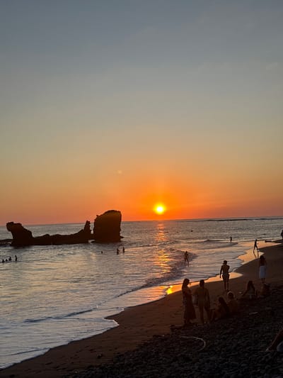 A vibrant sunset illuminates a beach scene. The sun casts a golden glow over the ocean, with silhouettes of people enjoying the water's edge. Rocks jut out from the sea, adding a dramatic element to the landscape. The sky transitions from blue to orange, mirroring the sun's radiant hues. The scene is captured in a vertical composition, accentuating the height of the sky and the depth of the ocean.