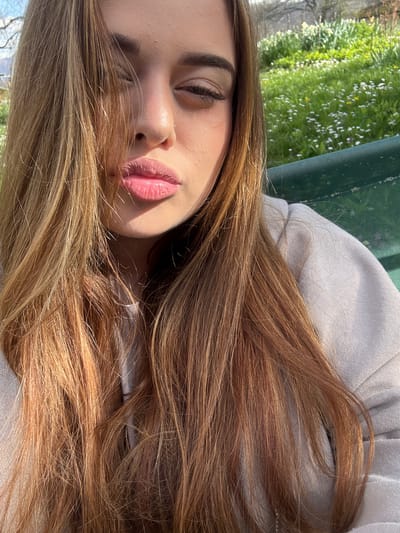 A young woman with long, light brown hair lies with her head tilted back, eyes partially closed, and lips slightly parted. She is wearing a light-colored hooded sweatshirt. The background features green foliage with small white flowers, suggesting a park or garden. The location is speculated to be Gorno Trape, Bulgaria.