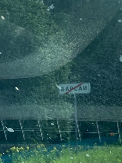 The image shows a sign with Russian text that reads "БАРСАИ" (Barsai) with a red diagonal line through it, indicating the end of a settlement or zone. The sign is mounted on a pole and is positioned next to a road or pathway, with a blurred background of greenery and what appears to be a fence or barrier. There are also some specks of light or dirt visible on the surface in the foreground, possibly a windshield. The overall scene is outdoors, with a focus on the sign marking the boundary of the settlement.