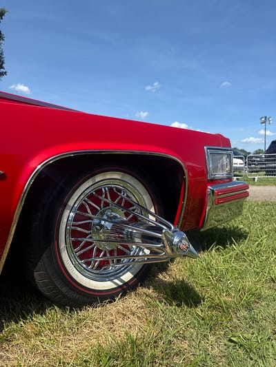 A low-angle, close-up shot captures the front quarter panel of a red Cadillac, showcasing its chrome details and wire wheels with whitewall tires. The car is parked on a grassy area, with a bright blue sky and scattered clouds overhead. In the background, out-of-focus elements suggest an outdoor event or gathering, with metal fencing, light poles, and other vehicles visible. The scene is devoid of any people. The location is Fairburn, United States.