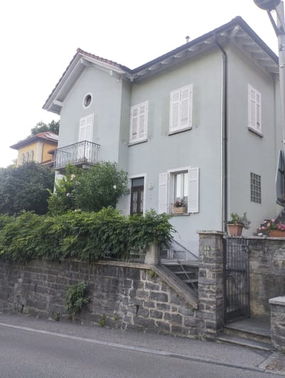 The image shows a light blue, two-story house with a red-tiled roof, situated in Orselina, Switzerland. The house has white shutters on its windows. A balcony with a decorative wrought-iron railing is visible on the second floor. In front of the house, there is a stone wall topped with a dense hedge of green bushes. A metal gate with a stone pillar stands to the right of the hedge, leading to a set of outdoor stairs. Potted plants with pink flowers are placed on the steps and a stone ledge. The house is bordered by a paved street in the foreground. The sky is overcast and bright.