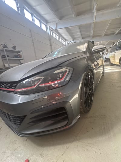 In a garage located in Schkopau, Germany, a dark gray Volkswagen Golf GTI is parked. The car is positioned at an angle, with its front left side prominently displayed. The vehicle appears dusty and possibly undergoing maintenance or repair, as indicated by the lighting and the overall environment which suggests a workshop or storage space.

The garage interior is characterized by concrete walls and a ceiling with exposed beams and fluorescent lighting fixtures. Large windows line the upper section of the wall on the left, allowing natural light to enter. Metal shelving units are visible against the back wall, holding various items.

The Golf GTI itself shows distinctive features of the GTI model, including the red accent stripe across the grille and headlights. The car is fitted with aftermarket black alloy wheels. In the background, a white van is partially visible, suggesting other vehicles might also be present in the garage. The floor is concrete, with some scuff marks and debris. The image is taken from a low-angle perspective, emphasizing the car's front end and wheel.