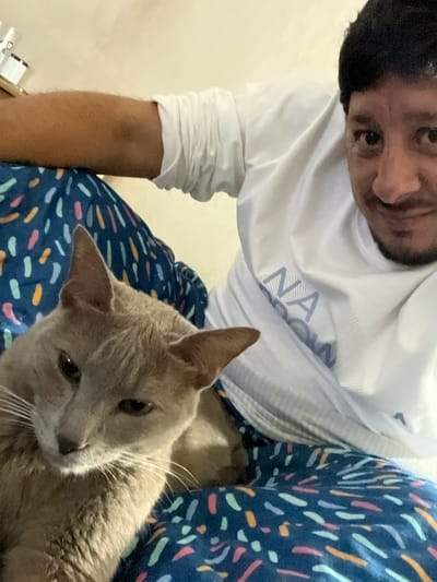 A close-up shot depicts a man and a cat. The man, with dark hair, is wearing a white long-sleeved shirt. He is looking down at the camera. The cat, light brown in color, is sitting on the man's lap. The man is wearing patterned pajama pants. The location cannot be determined from the image.