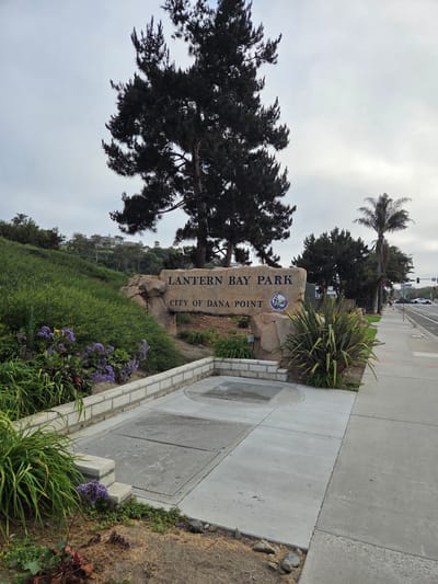 The image shows the entrance sign for Lantern Bay Park in Dana Point, California, USA. The sign, made of stone, reads "LANTERN BAY PARK" and "CITY OF DANA POINT". A circular emblem with a lighthouse and the text "DANA POINT CALIFORNIA 1989" is to the right of the park name. The sign is situated on a landscaped hillside with lush green vegetation and purple flowers in the foreground. To the right of the sign is a sidewalk and a street with traffic, indicating an urban or semi-urban environment. Tall evergreen trees and palm trees are visible, along with buildings in the background on the hillside. The sky is overcast.