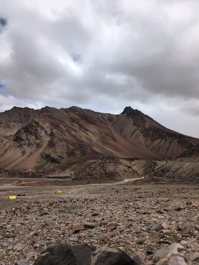 The image depicts a vast, rocky terrain with a dramatic mountain range dominating the background. The sky is overcast with heavy, grey clouds. The mountains are rugged and appear to be made of earth and stone, with varying shades of brown, tan, and grey. Streaks of darker rock and some sparse vegetation are visible on their slopes. A winding dirt road is discernible, leading through the lower slopes of the mountains. In the middle ground, several yellow tents are set up on the rocky ground, suggesting a temporary encampment. The foreground is covered in loose rocks and pebbles of various sizes. The scene appears to be a remote, natural landscape, possibly in an arid or high-altitude region. No people are visible in the image. The location is identified as Killingsarai, India.