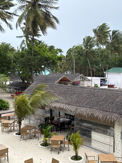 This image shows a tropical beach resort area in Dhigurah, Maldives. The scene is dominated by palm trees swaying in the wind, with a cloudy sky overhead. In the foreground, there is an outdoor seating area with thatched-roof structures and numerous tables and chairs set up on a sandy surface. Some of these tables appear to be made of wood, with metal chair frames. Further back, more thatched-roof buildings are visible, along with lush green vegetation and glimpses of a turquoise ocean. The architecture suggests a laid-back, tropical resort ambiance. There are no people visible in the image, and no apparent actions or interactions taking place. The environment appears somewhat windswept, indicated by the movement of the palm fronds.