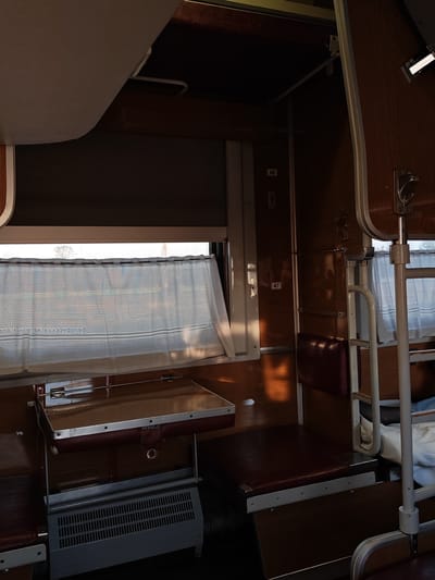 The image depicts the interior of a train compartment. The walls are paneled with wood. There is a window with a curtain. A folding table is in front of the window, with a heater underneath. On the right, there's a bunk bed with a ladder and a partially visible pillow and blanket. Numbers "48" and "47" are visible on the wall, suggesting the compartment designation. The location is within Мажэйкаўскі сельскі Савет, Belarus.