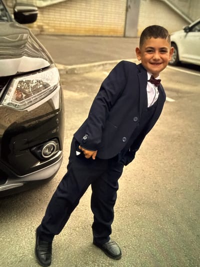 A young boy, appearing to be around 8 years old, is pictured outdoors, leaning towards a dark-colored SUV parked on a paved surface. The boy is dressed in a dark navy blue suit, complete with a vest and a burgundy bowtie. He is smiling and looking towards the camera. The SUV's headlights and front bumper are visible on the left side of the frame. In the background, a white car is partially visible, and a brick wall can be seen. The scene appears to be a street or parking lot in Artik, Armenia.