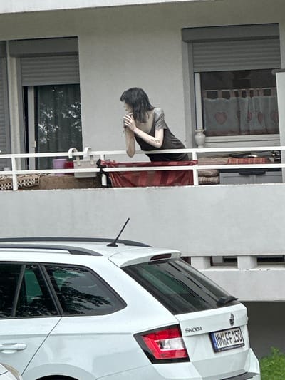 The image depicts a residential balcony with a mannequin positioned on it, appearing to lean over the railing and look outwards. The mannequin has dark, short hair and is wearing a dark-colored top. In the background, a window with white curtains adorned with red hearts is visible. On the balcony, various items are present, including what appear to be cushions or folded blankets, a purple bucket, and a white rectangular container. Below the balcony, the rear portion of a white Skoda car is visible, with its license plate reading "MM FF 150." The car is parked in front of the building. The overall scene suggests an outdoor urban or suburban residential setting.