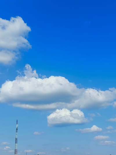 This media file captures a bright, clear day in Pukhivka, Ukraine, with a vast expanse of blue sky dominating the frame. Fluffy white cumulus clouds are scattered across the sky, some large and billowy, others smaller and more wispy. In the lower portion of the image, a tall, lattice-like communication tower stands prominently against the sky. The tower has a distinctive alternating pattern of white and dark blue or black sections. Below the tower, a sliver of land is visible, with faint hints of vegetation or structures. The overall impression is one of a tranquil, sunny day, likely in the daytime, given the bright blue sky and the vivid white of the clouds. There are no people or explicit activities visible, suggesting a focus on the landscape and the man-made structure within it. The clear weather and expansive sky contribute to a feeling of openness and peace. No visible text is present in the image.