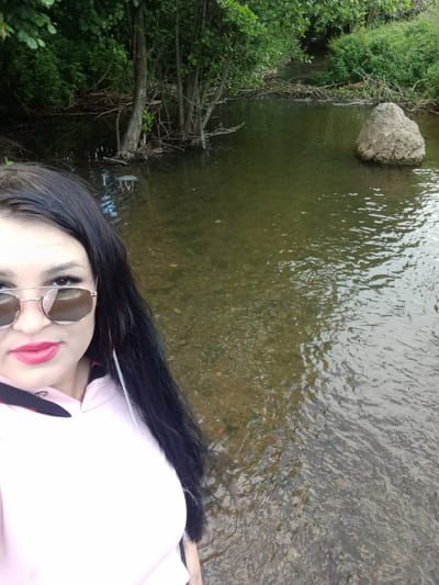 The media file is a self-portrait of a woman taken outdoors. The woman is wearing sunglasses and a pink top with a black collar. She has long black hair and bright pink lipstick. She is standing in shallow water, possibly a river or stream, with trees and foliage visible in the background. A large rock is submerged in the water to her right. The lighting suggests it is daytime. The overall mood appears casual and relaxed. There is no visible text in the media file.