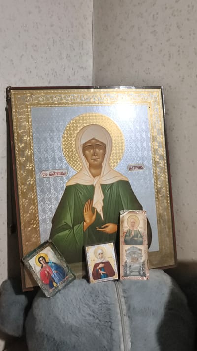 This image depicts a collection of religious icons displayed in a domestic setting. The primary icon, a large, framed depiction of a woman with a halo, is the central focus. It is positioned in the corner of a room with textured, light-colored wallpaper. To the right of this large icon, and slightly in front of it, are two smaller icons. One is a rectangular icon with two figures and a halo. Another is a smaller, square icon depicting a single female figure with a halo, and to its right is a tall, narrow icon with two depictions of religious figures. The icons are resting on a soft, grey, fuzzy surface, possibly a piece of furniture or fabric. The lighting appears to be artificial and casts shadows, indicating an indoor environment. The presence of multiple icons suggests a devotional space. However, there is no information in the image that can confirm its location in the city of Мирзо Турсунзода, Tajikistan.