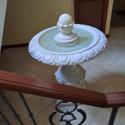 A white marble fountain with a carved pineapple-like top is situated indoors on a tiled floor. The fountain is filled with clear water. A wooden handrail and dark metal railing are in the foreground, obscuring part of the fountain and the floor. The background includes a wall with a lattice-like wooden panel and another wall painted in a light yellow hue. The location is identified as Tairove, Ukraine.