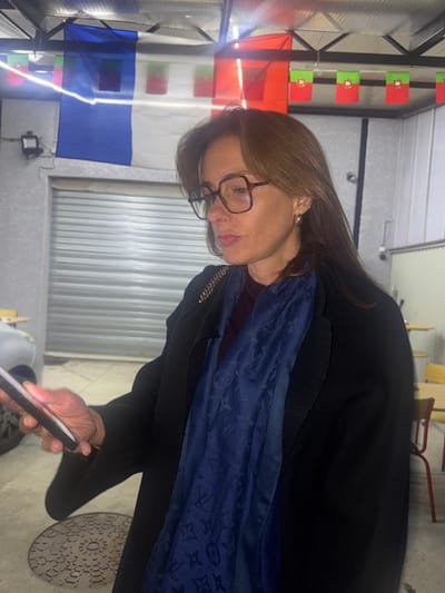 The scene depicts a woman in an indoor or sheltered outdoor setting in Décines-Charpieu, France. The woman, appearing middle-aged, has medium-length brown hair and is wearing dark-framed eyeglasses and small gold hoop earrings. She is dressed in a dark coat or jacket over a dark red garment, with a dark blue scarf, patterned with what appears to be a Louis Vuitton monogram, draped around her neck. A gold-colored chain strap is visible on her right shoulder. She is holding a smartphone in her left hand, looking down at its illuminated screen and seemingly interacting with it; a ring is visible on her left ring finger.
The background features a large French flag (blue, white, red vertical stripes) hanging prominently from an overhead structure, likely a corrugated metal roof. To the right of the French flag, a string of smaller Portuguese flags (red and green with the national emblem) is also displayed. A light-colored wall with horizontal ridges, possibly corrugated metal siding, is behind the flags. Below the French flag, a closed metal roller shutter is visible, suggesting a secured entrance or garage. Partially visible on the far left is the front portion of a light-colored vehicle. In the blurred background on the right, there are yellow-topped tables with chairs featuring red legs, indicating a casual seating area. On the concrete ground directly below the woman's left hand, a circular metal manhole cover with an intricate pattern is present. The artificial lighting source overhead creates a glare in the image. The flags suggest a connection to both French and Portuguese culture.