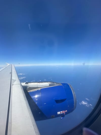 The image is an aerial view from an airplane window, showing a blue airplane engine and a portion of the wing against a vast expanse of the ocean and a clear blue sky. The horizon is visible, with scattered clouds low on the water's surface. The scene depicts a calm flight over the sea, with no indication of the city of Viehhausen, Austria.