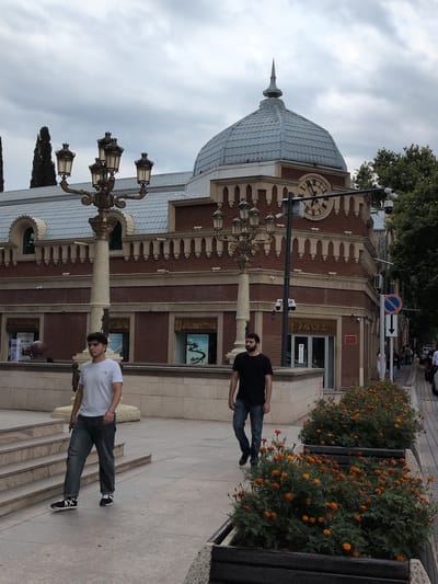 The image depicts a street scene in Ganja, Azerbaijan. In the foreground, two men are walking on a paved area. The man on the left is wearing a white t-shirt and dark jeans, while the man on the right is wearing a black t-shirt and dark jeans. They are walking in the same direction, away from the viewer.

Behind the men is a building with a red brick facade and a domed roof, characteristic of some architecture found in the city. A large clock is visible on the side of the building. A street lamp with multiple fixtures stands to the left of the building. Flower planters with orange marigolds are positioned in the foreground along the sidewalk.

The sky is overcast, suggesting a cloudy day. The overall scene is that of an urban environment, possibly a public square or street with pedestrian traffic and commercial establishments. The building's architecture and the presence of a clock tower are notable features.