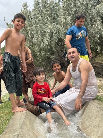 In Kan Mehtarzai, Pakistan, a group of males, including at least three children and two adults, are seen in what appears to be an outdoor water channel or ditch. The environment suggests a natural setting with a large tree with light-colored foliage dominating the background. The main subjects are positioned in and around the water channel.

In the foreground, a young boy in a red long-sleeved shirt and dark pants is seated, with water splashing around his legs. He is smiling and looking towards the camera. To his left, a young boy with no shirt on and dark, baggy pants stands with a wide, open-mouthed smile, suggesting excitement. Another adult male, wearing a white tank top and light-colored, loose-fitting pants, is seated in the water channel to the right of the children, looking directly at the camera.

Behind these individuals, another young boy, shirtless, is visible. Further back, an adult male in a blue t-shirt and yellow shorts stands, looking down. Another adult in a brown, traditional garment stands partially obscured by the others. The water in the channel is actively flowing, creating splashes and ripples around the individuals. The ground around the channel appears to be covered in grass.