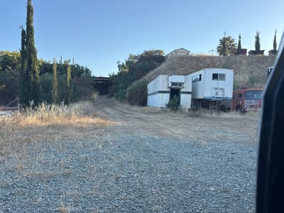 The image depicts an outdoor scene at what appears to be a junkyard or an abandoned property. The setting is rural, with dry, yellowed grass and gravel covering the ground. In the foreground, there is a patch of gray gravel. Behind it, a dirt track leads uphill.
Dominating the middle ground are two white, rectangular structures that resemble portable offices or storage containers. One has an open doorway with some greenery growing around its base. Next to these structures, a vintage red truck is parked. The truck appears old and possibly in disrepair.
Further up the hill, a single-story building with a light-colored roof and walls is visible, nestled amongst dry grass and trees. The landscape is hilly, with slopes covered in vegetation that appears dry and sparse in places. Several tall, slender cypress trees stand to the left of the image, adding verticality to the scene. In the background, a dark structure, possibly a bridge or an overpass, is visible spanning a gap. The sky is clear and blue, suggesting it is daytime with fair weather. The overall impression of the scene is one of disuse and neglect. There are no people visible in the image. The time of day appears to be late afternoon or early morning, given the angle of the sunlight casting shadows. There is no visible text in the image.